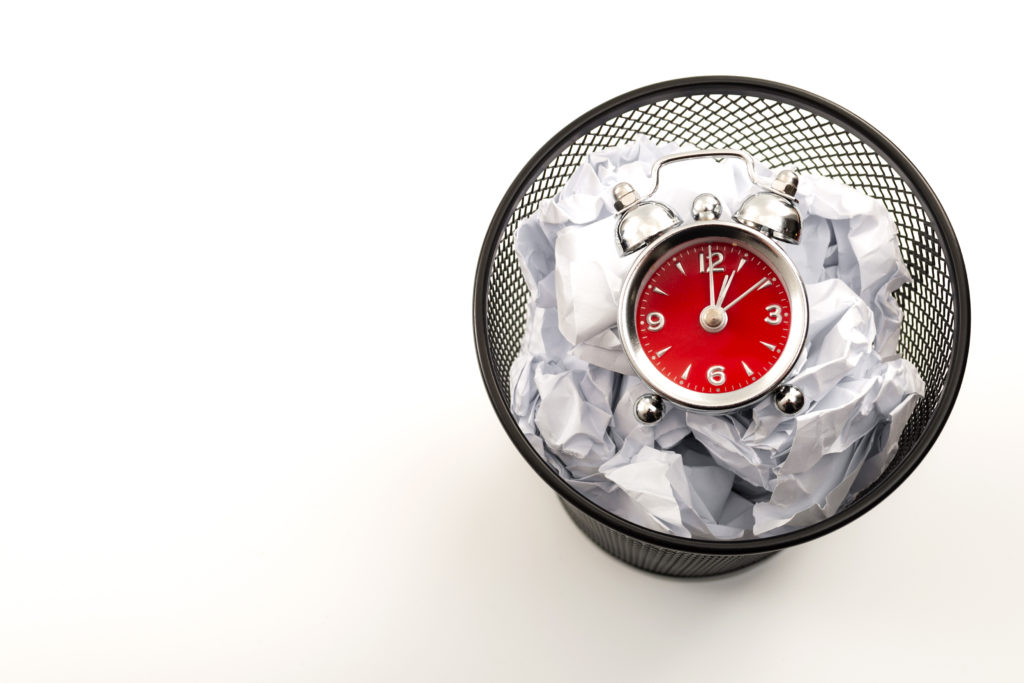 Procrastination is the enemy of productivity and stop wasting time concept theme with a clock in a black waste basket or rubbish bin next to crumpled paper isolated on white background with copyspace Red watch in trash bin along with papers